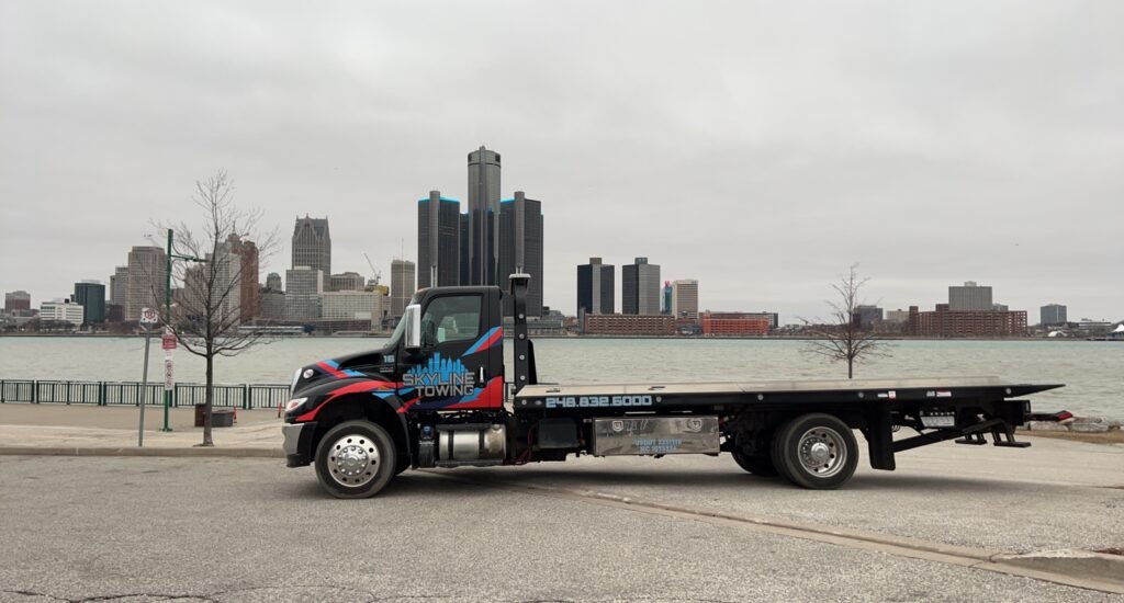 Empty black Skyline Towing International flatbed parked on the riverfront with the Detroit skyline and Renaissance Center across the river